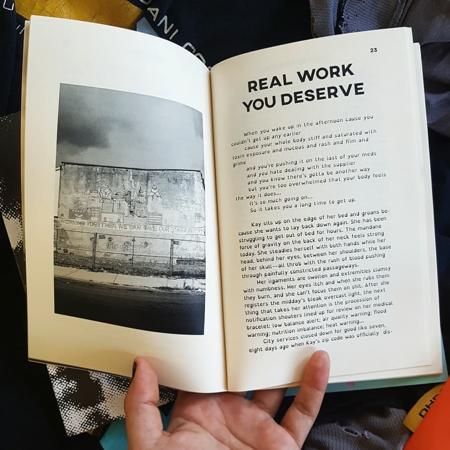 Im holding open the zine. A b/w image of a building with a mural on it is on the left. The right has a story titled REAL WORK YOU DESERVE.