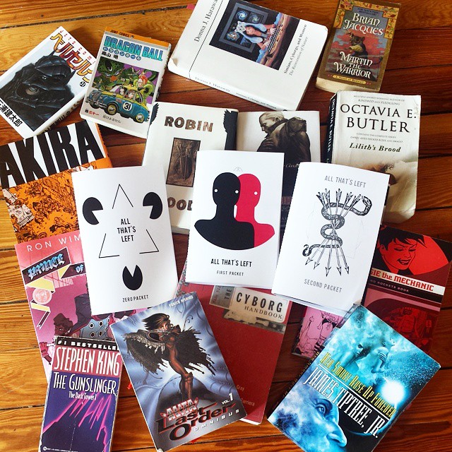 A wood floor strewn with books and comics, with my ATL zines in the center. Some of the comics are AKIRA, BERSERK, Dragon Ball, Love and Rockets, Battle Angel Alita. Some of the books are The Cyborg's Handbook, the Gunslinger, Her Smoke Rose Up Forever by James Tiptree Jr, Octavia Butler, Robin Hood, the Redwall series, and a Ghost in the Shell novel in English.