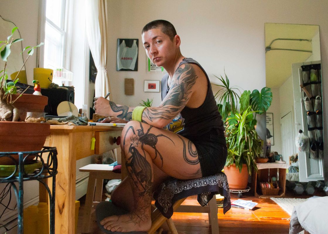 The author sits in profile at their desk, looking at the camera. They are white mixed, with arms and leg etched with tattoos of flames and spiders and snake tails. Happy houseplants are all around the room.