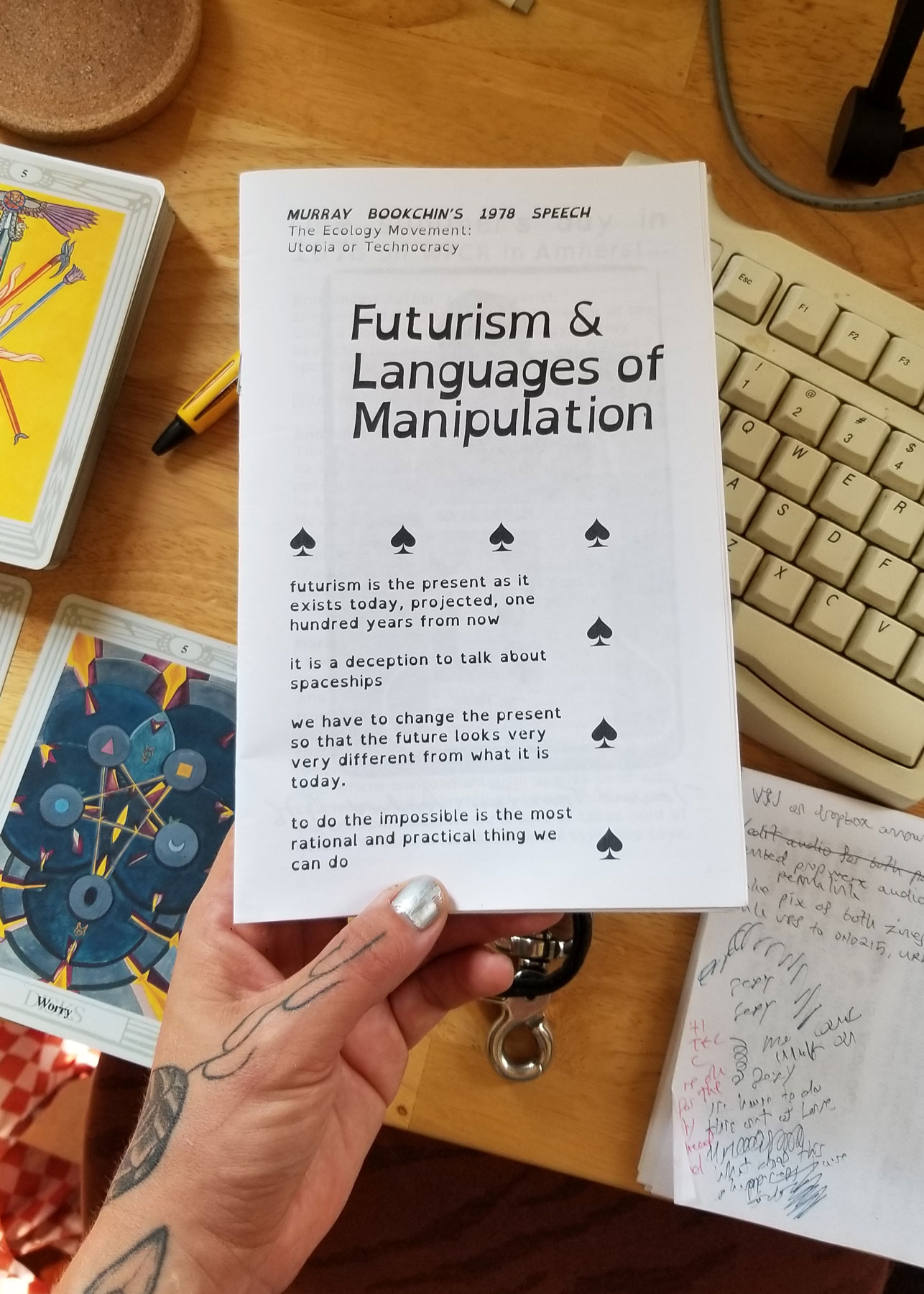 My left hands hold the booklet zine , with my keyboard and some tarot cards on my desk in the background. The zine says FUTURISM & LANGUAGES OF MANIPULATION