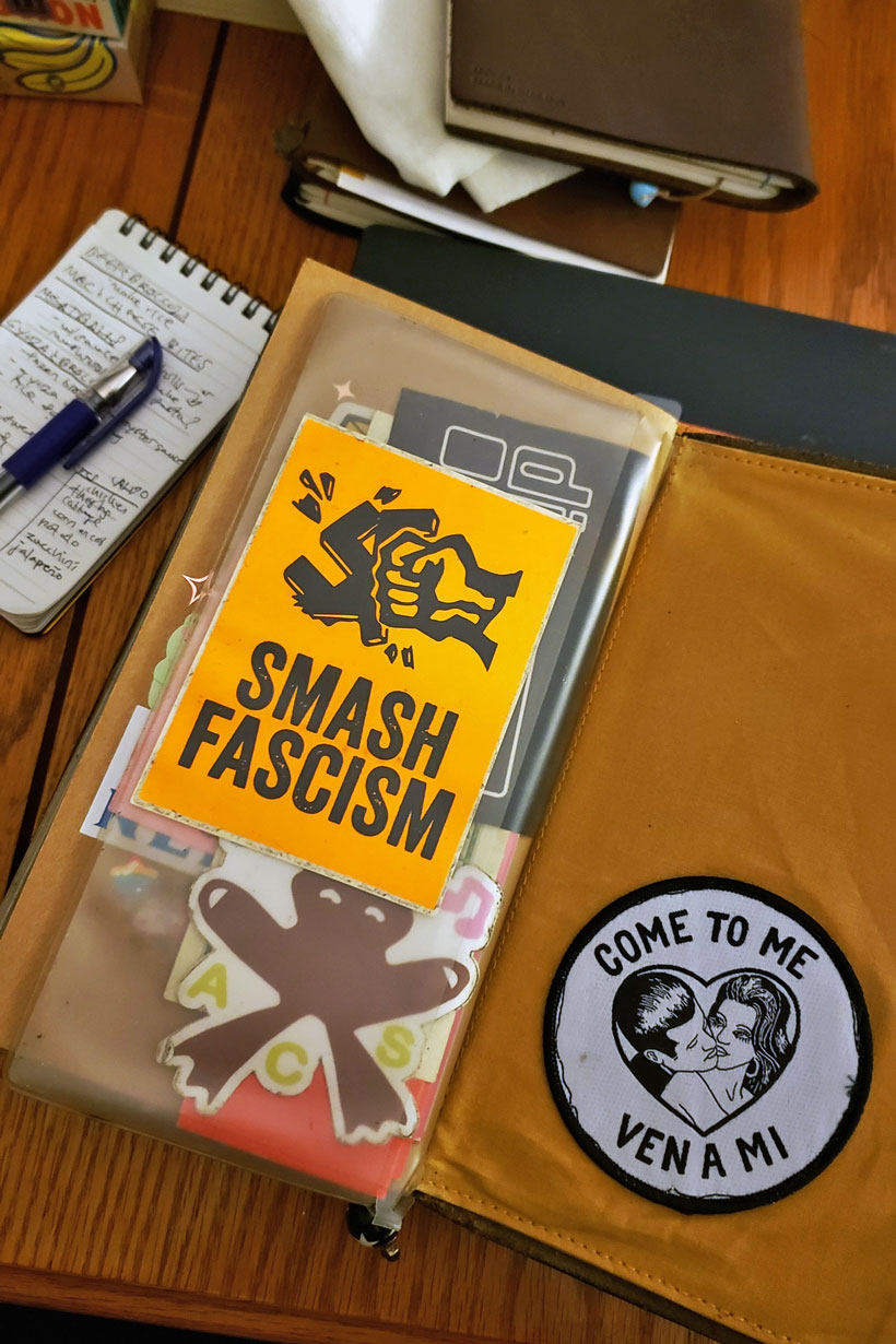 a wood table full of journals and memo pads. a sticker pouch sits on top next to a canvas pouch w/a patch that says come to me ven a mi [[shout out Tamara Santibañez]]; the biggest sticker in the case says smash fascism